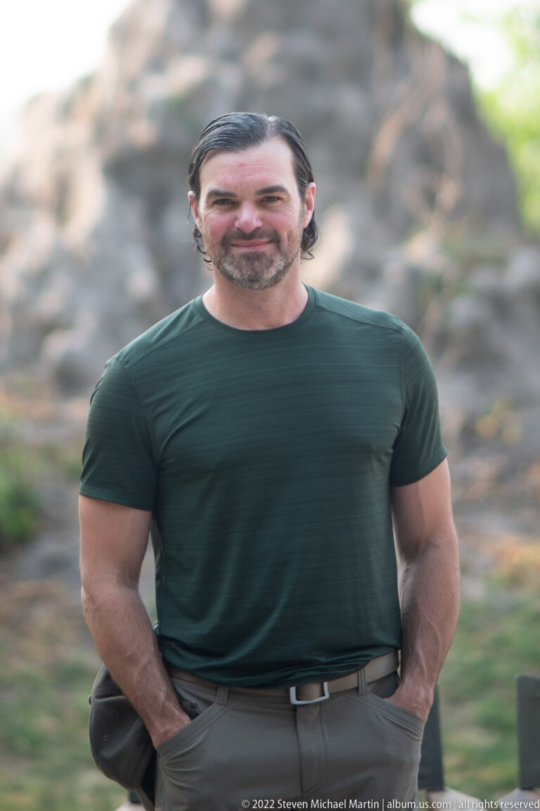 Close portrait of a safari traveler in a dark green shirt with a large termite mound blurred behind.