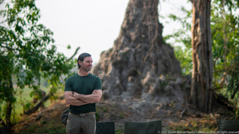 Safari traveler standing with arms folded near a large termite mound framed by trees and camp chairs.