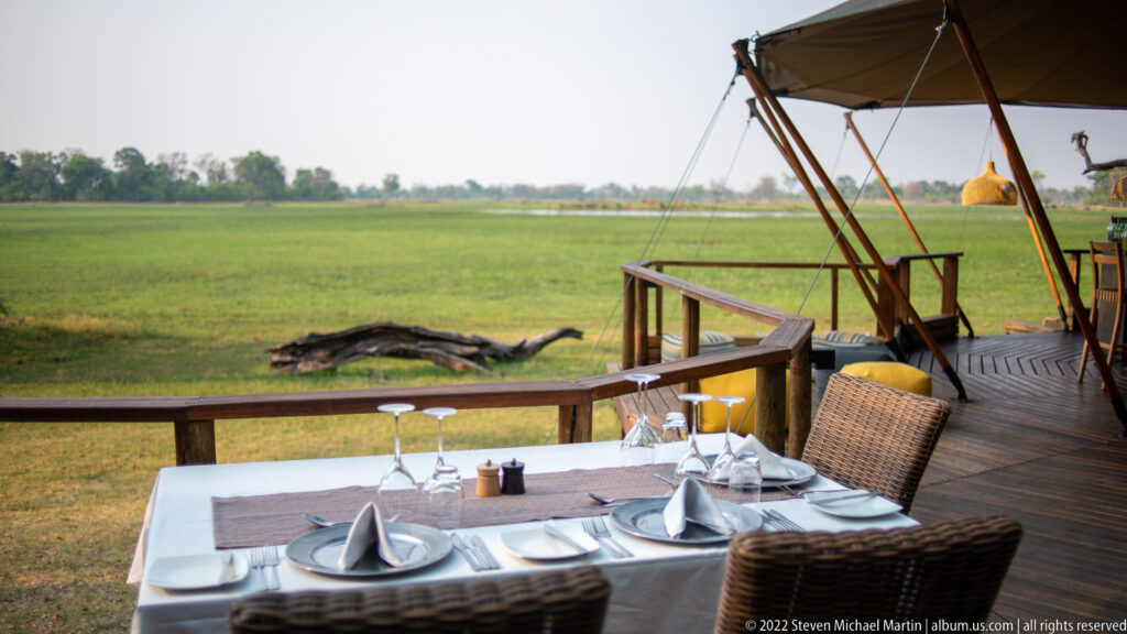 Set dining table with plates, folded napkins, and wine glasses on a wooden terrace overlooking a green floodplain.