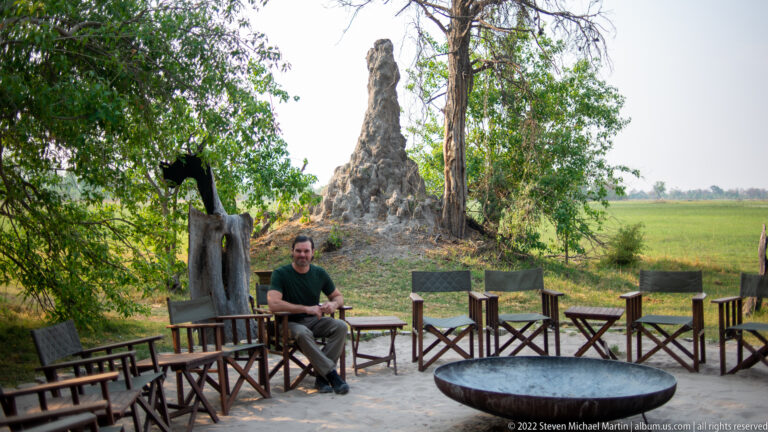 Safari chairs arranged around a metal fire bowl on sand, with a large termite mound and trees behind.