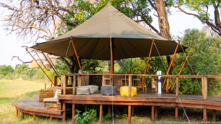Raised open-sided safari pavilion with canvas roof, timber poles, wooden deck, railings, chairs, and tables among trees.
