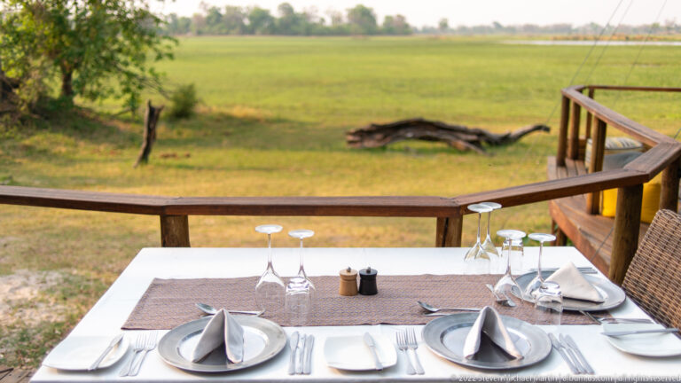 Close view of a formal table setting with gray napkins and glasses beside a railing overlooking green floodplain.