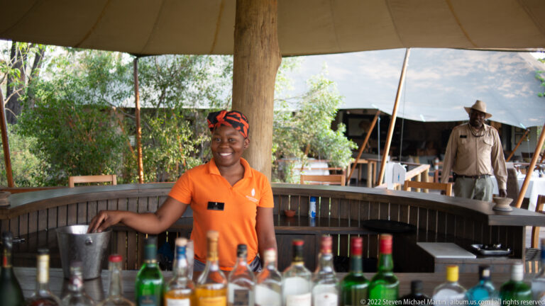 Smiling camp staff member in orange standing behind an open-air bar lined with bottles under a canvas roof.