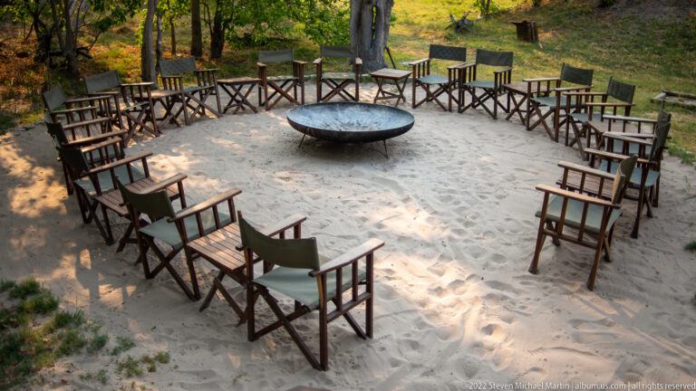 Elevated view of wooden safari chairs arranged in a circle around a large metal fire bowl on pale sand.