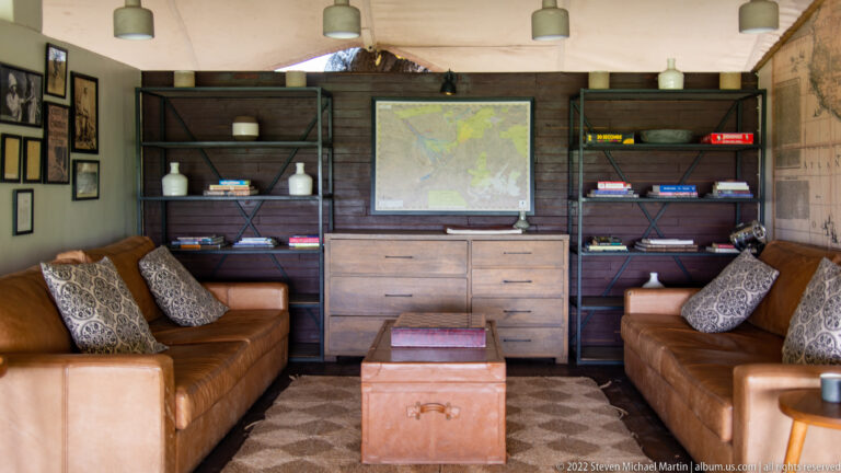 Safari lodge lounge with facing leather sofas, central trunk, dark bookshelves, and a large wall map.