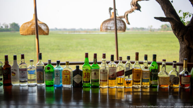 Row of assorted clear, green, and amber bottles on an open-air bar with green floodplain visible behind.
