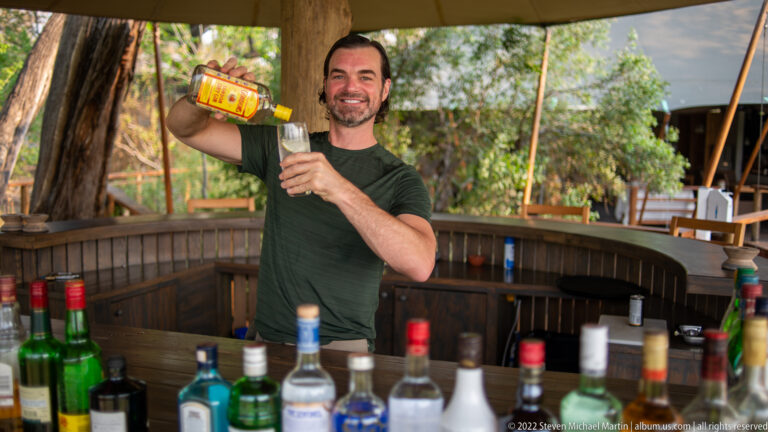 Safari traveler smiling while pouring a drink into a tall glass behind an open-air bar lined with bottles.