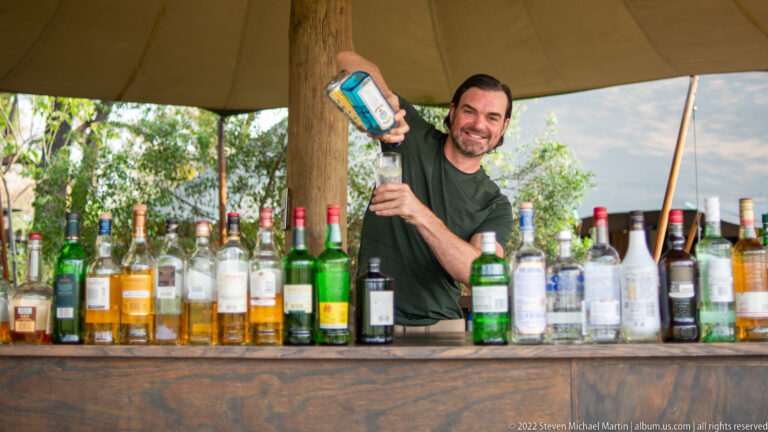 Safari traveler behind a wide wooden bar raising a blue bottle over a tall glass amid many bottles.