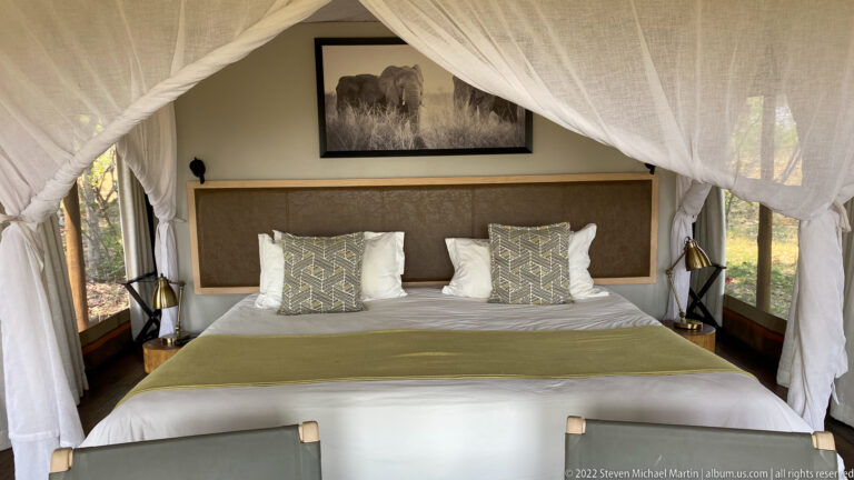 Large safari-lodge bed beneath white mosquito netting with neutral linens, patterned pillows, lamps, and wildlife artwork.