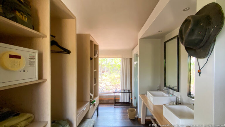 Open-plan safari-lodge dressing area with shelving, safe, hanging space, twin sinks, mirrors, and a bright doorway beyond.