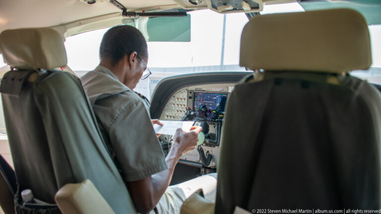 Pilot seated at the instrument panel of a small aircraft, viewed from behind between the front cockpit seats.