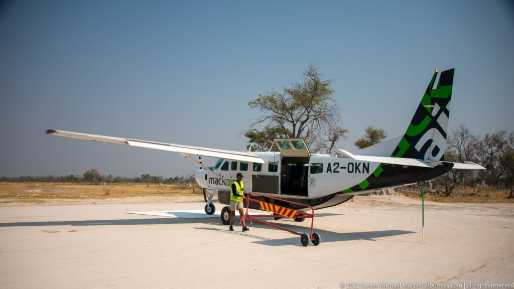 High-wing Mack Air light aircraft parked on a small airstrip with its cabin door open and a ground worker beside it.