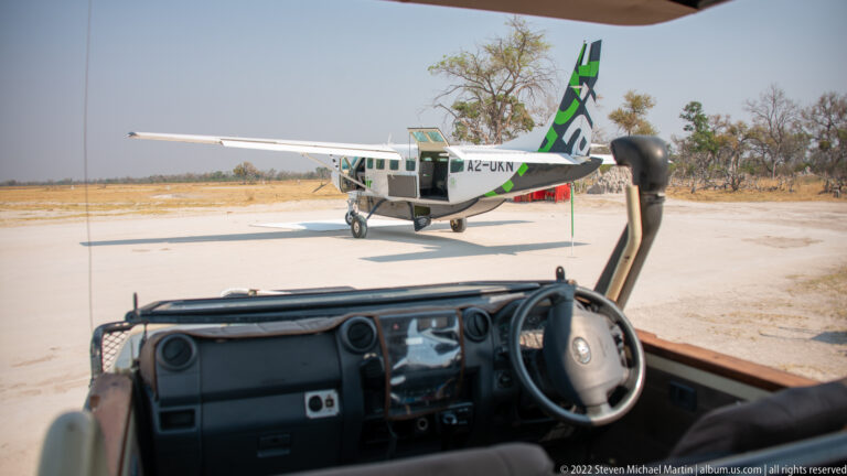 View from an open safari vehicle toward a parked Mack Air light aircraft on a remote Okavango airstrip.