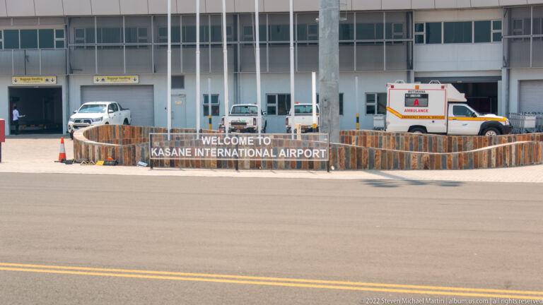 Kasane International Airport terminal seen across the apron, with a welcome sign, service vehicles, and an ambulance.