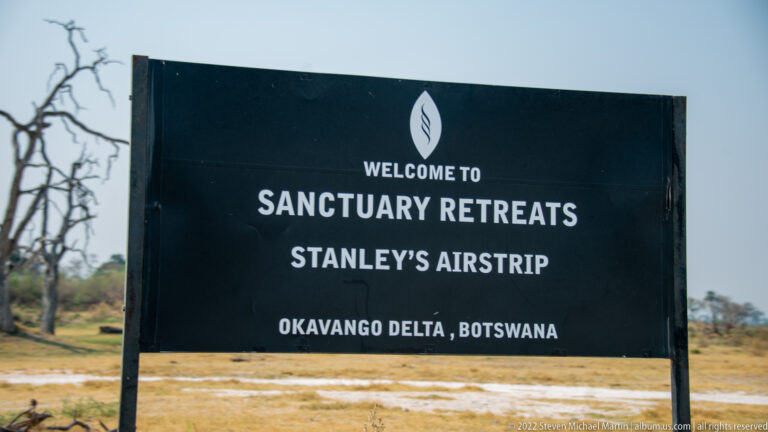 Black sign reading 'Welcome to Sanctuary Retreats Stanley’s Airstrip, Okavango Delta, Botswana' beside a sandy airstrip.