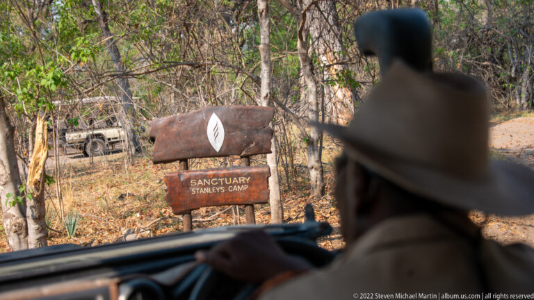 Safari driver passing a wooden Sanctuary Stanley’s Camp sign on a sandy track through woodland.