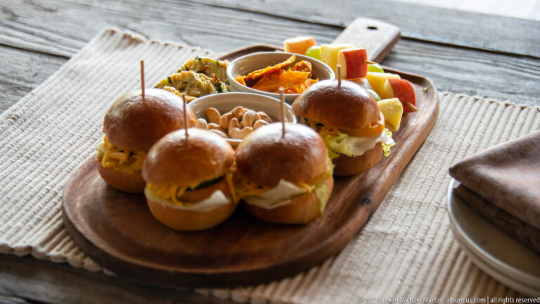 Wooden board holding several small savory sliders, nuts, dips, and colorful snack portions on a woven table runner.