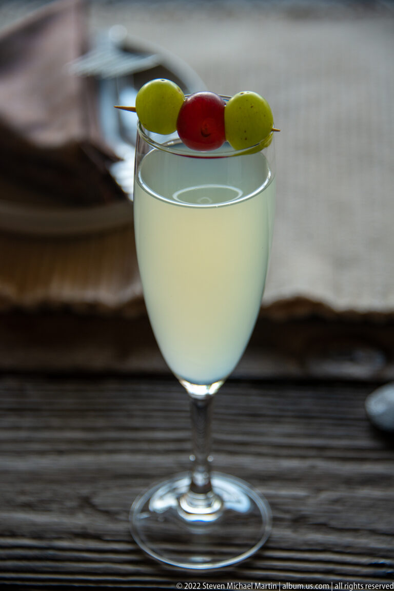 Pale drink in a rounded stemmed glass with red and green garnish pieces balanced across the rim.