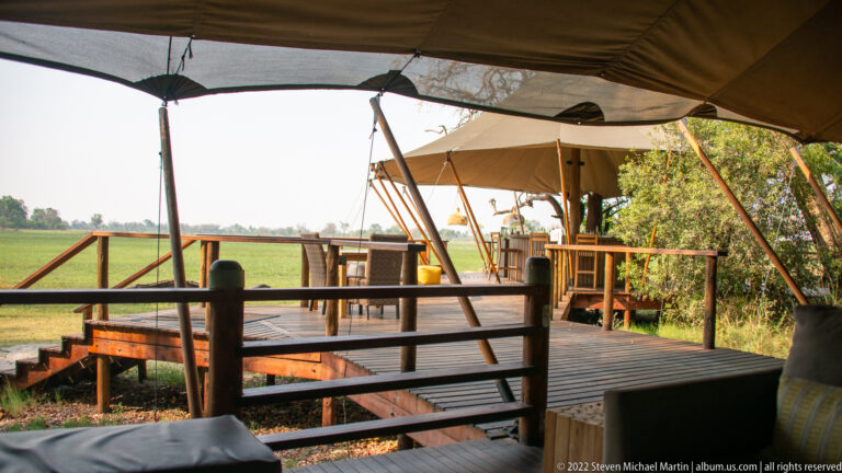 Raised wooden deck with open canvas lounge and dining structures overlooking a green Okavango floodplain.