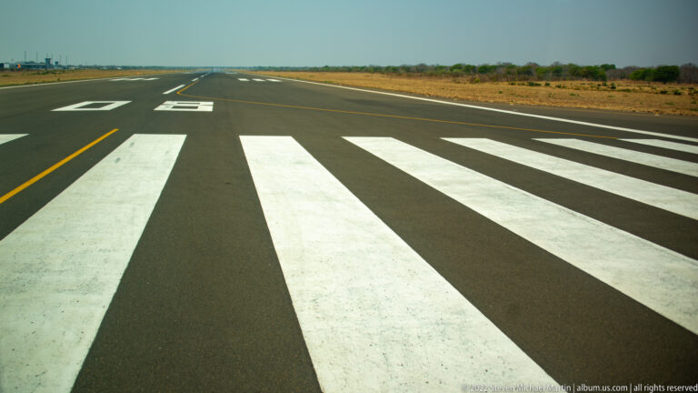 White runway threshold bars and runway numerals on dark pavement, with dry scrubland beyond the runway.