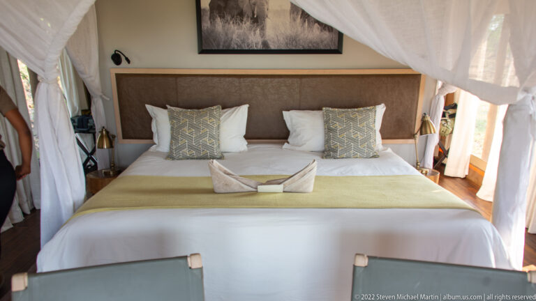 Large neatly made bed with patterned cushions and white mosquito netting inside a canvas safari tent.