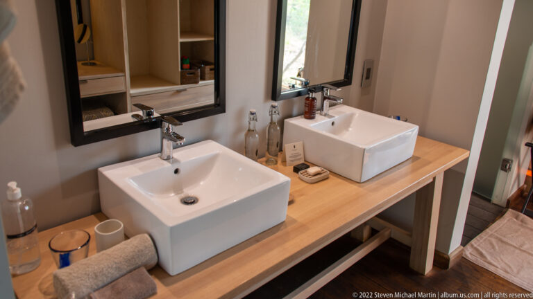 Two square white vessel sinks on a wooden vanity beneath dark-framed mirrors, with bottles, towels, and toiletries arranged around them.