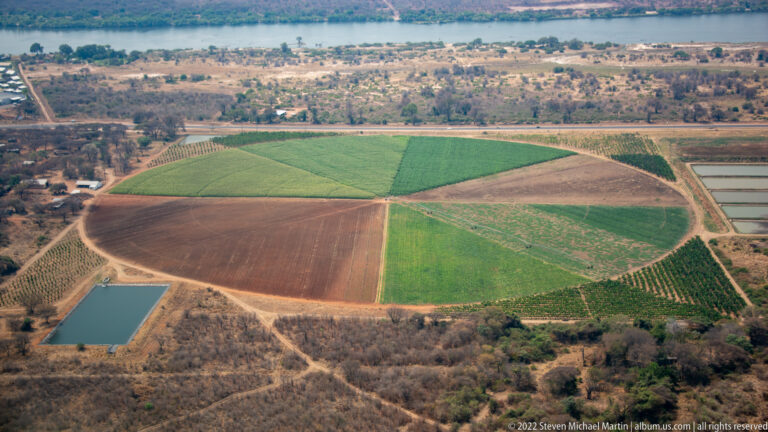 Aerial view of a large circular irrigated field, smaller farm plots, ponds, and surrounding dry bush.