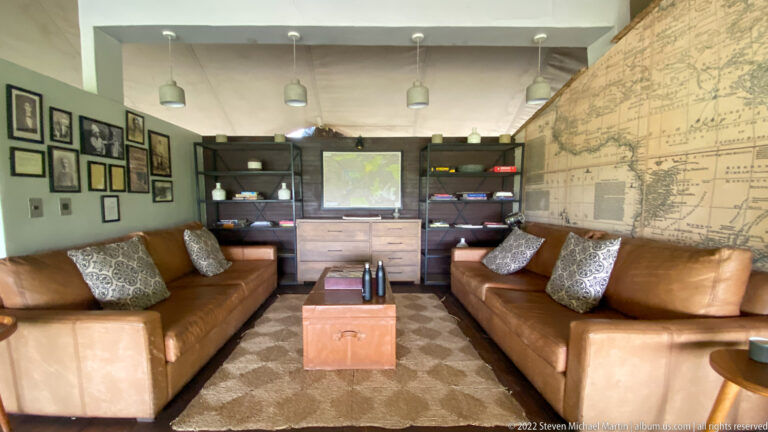 Safari camp lounge with two brown leather sofas, patterned cushions, bookshelves, framed pictures, and a large wall map.
