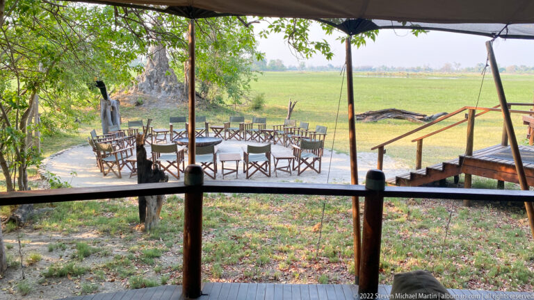 Outdoor dining tables and chairs arranged beneath trees beside a green Okavango floodplain, viewed from a raised deck.