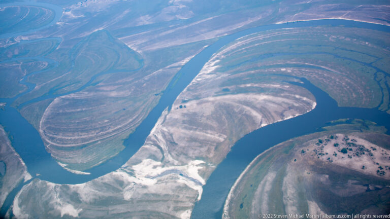 Aerial view of a dark winding channel forming tight meander bends through pale exposed sediment and floodplain.