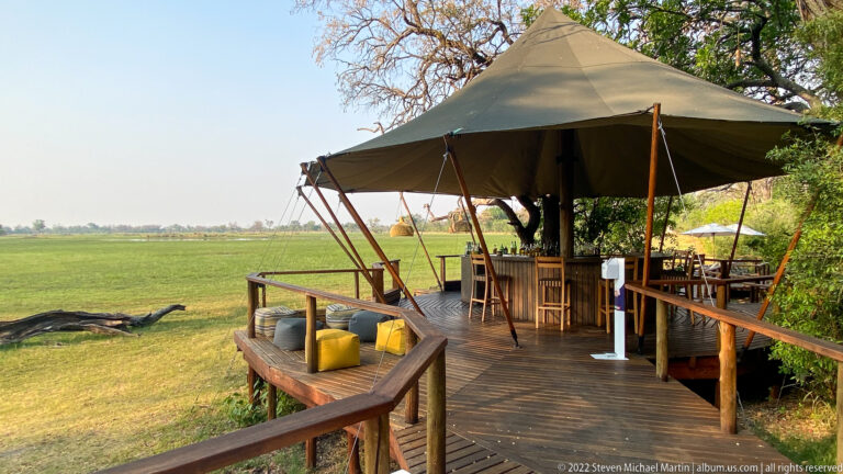 Canvas-roofed bar on a raised wooden deck with stools and seating overlooking the green Okavango floodplain.
