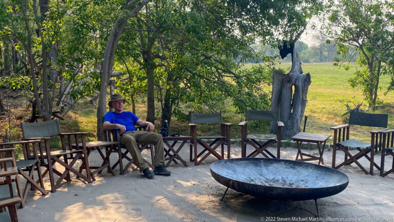 Guest seated among wooden camp chairs around a large metal fire bowl on sand beneath trees.