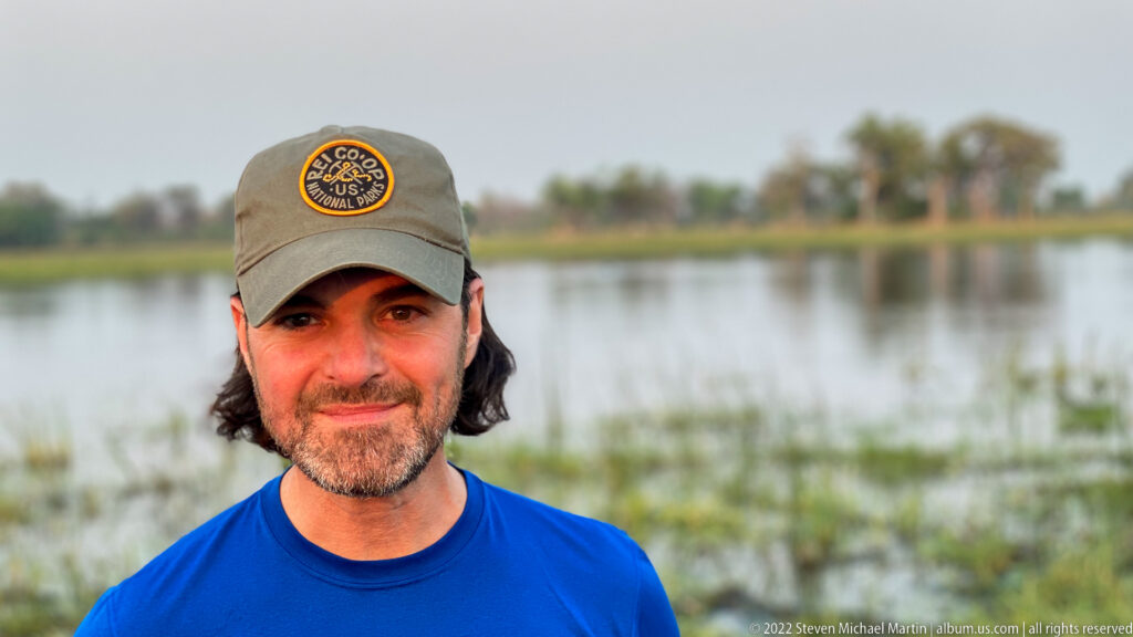 Close portrait of a safari guest in a blue shirt and olive cap, with shallow wetland water blurred behind.