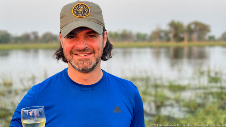 Smiling safari guest in a blue shirt and cap holding a stemmed drink beside blurred wetland water.