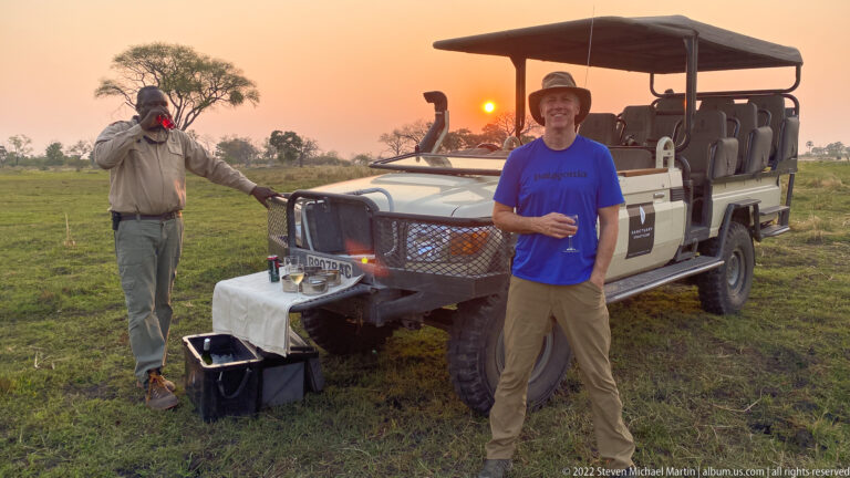Safari guide and guest standing beside an open game-drive vehicle at sunset, with drinks arranged on the front.