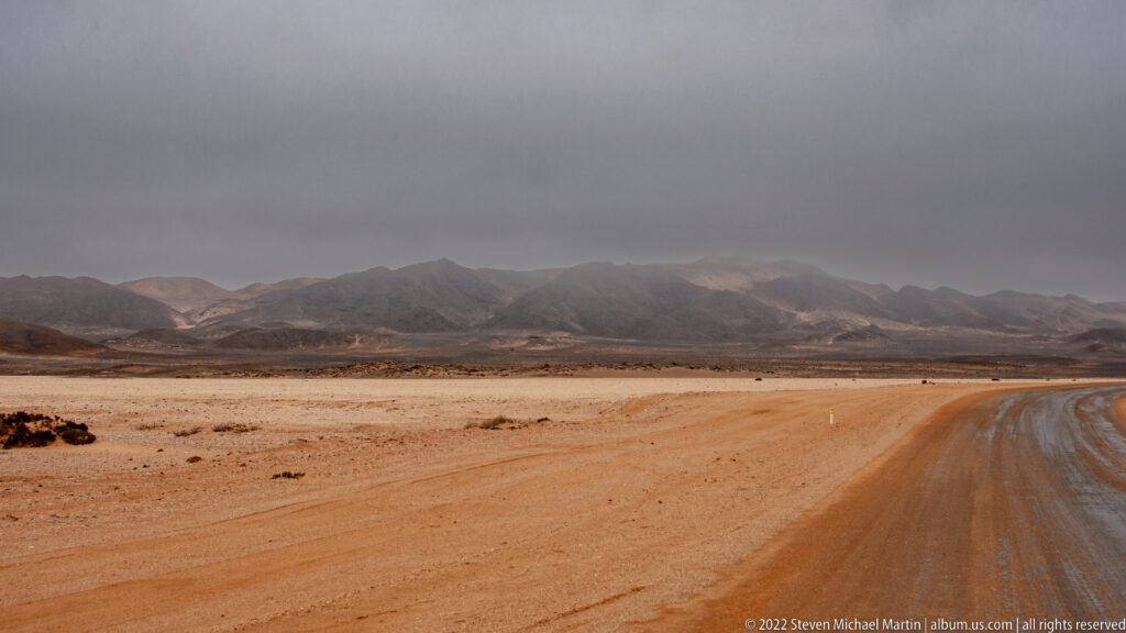 Unpaved desert road curving across a barren Namib plain toward dark mountains beneath low gray cloud.