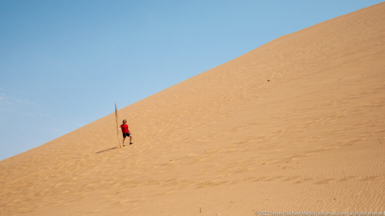 Red-shirted visitor crossing a steep orange dune face with footprints trailing across the sand.