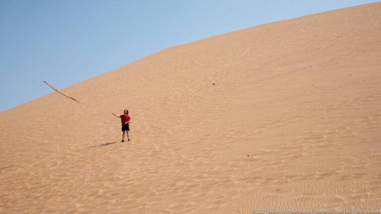 Person in red standing on a steep dune with one arm extended and a long pole angled across the sand.