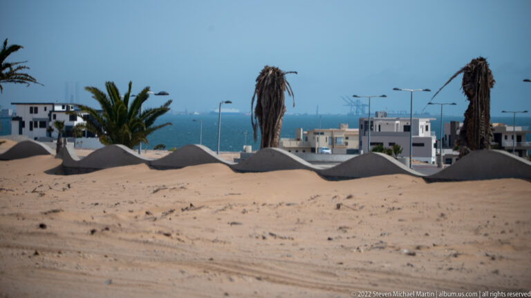 White coastal buildings and palms beyond windblown sand, with blue sea and faint port cranes in the distance.