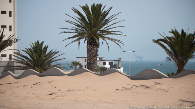 Tall palms behind a low wavy wall at the sandy edge of a coastal settlement, with harbor cranes beyond.