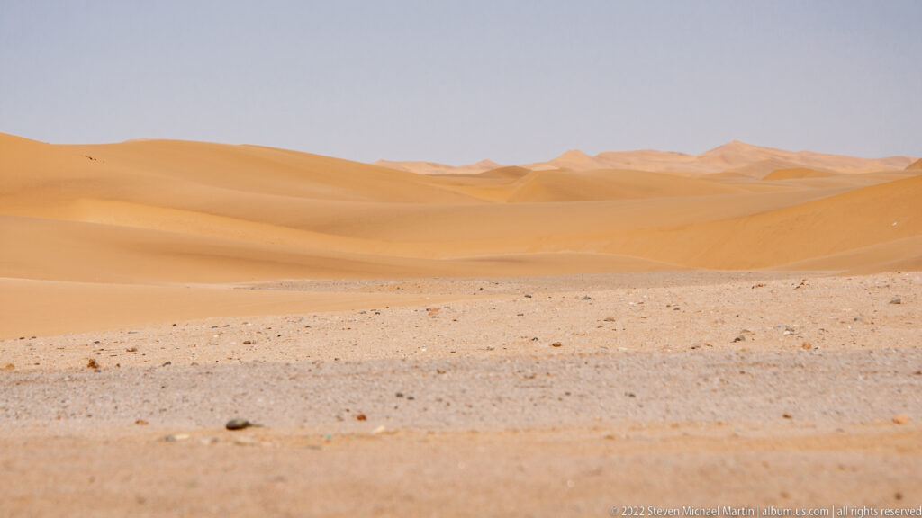 Layered orange sand dunes beyond a pale stony desert foreground under a hazy blue-gray sky.