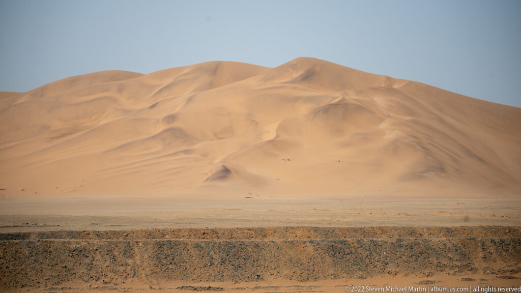 Massive sculpted orange dune complex rising from a flat dark gravel plain beneath a pale blue sky.