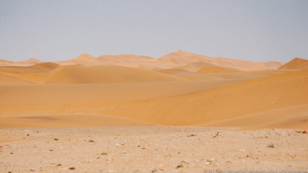 Sweeping orange dune ridges with high crests rising behind a narrow gravelly foreground in the Namib.