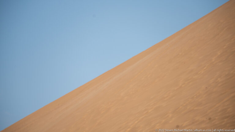 Steep orange sand dune slope forms a diagonal against an uninterrupted blue sky, with faint wind-textured surface markings.