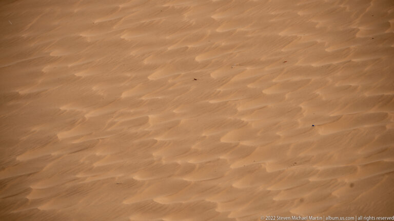 Close view of ochre dune sand patterned by wind-made ripples, shallow hollows, and scattered dark specks.