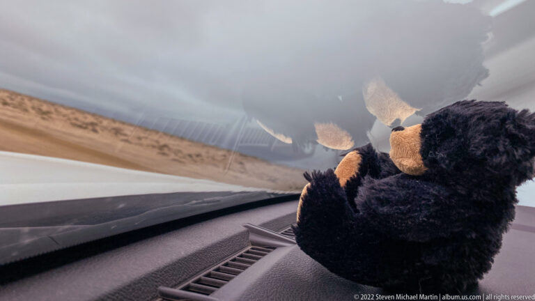Dark plush primate toy sits on a vehicle dashboard beside the windshield with sandy desert terrain visible outside.