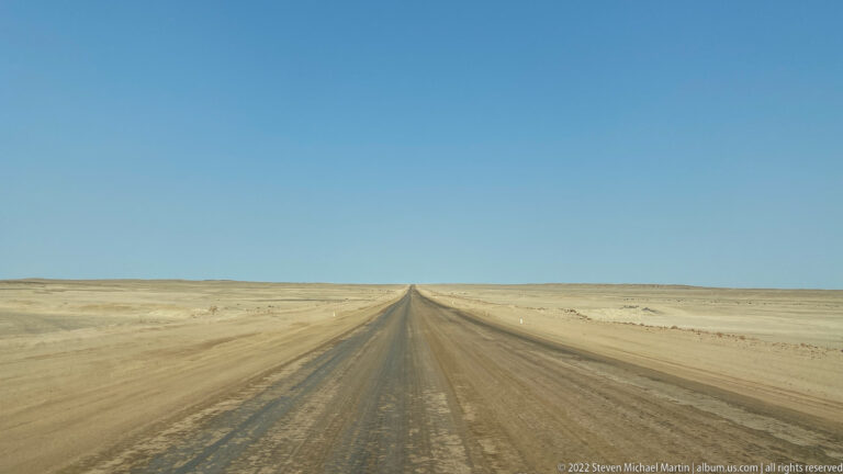Straight gravel road extends to the horizon across a flat, sparsely vegetated Namibian desert plain under a blue sky.