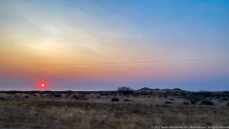Red-orange sun sets over dry scrubland with silhouetted bushes, distant low hills, and a thin utility line.