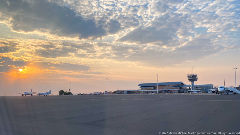 Airport apron at sunset with parked aircraft, low terminal buildings, light poles, and an air traffic control tower.