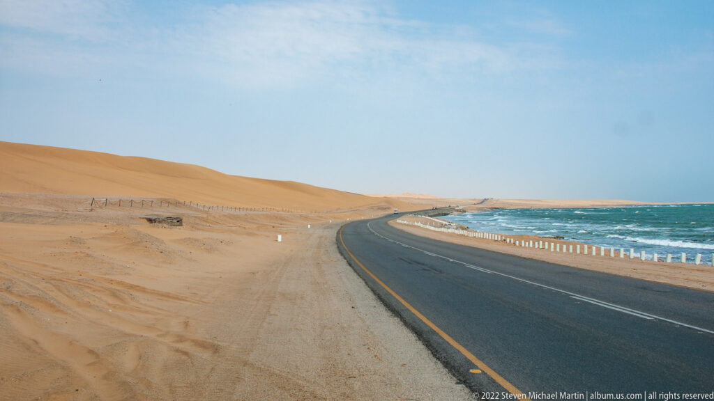 Paved highway curving between orange desert dunes on the left and blue ocean surf on the right.